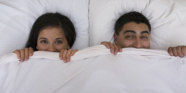 Multi-ethnic couple holding bed sheet over mouths